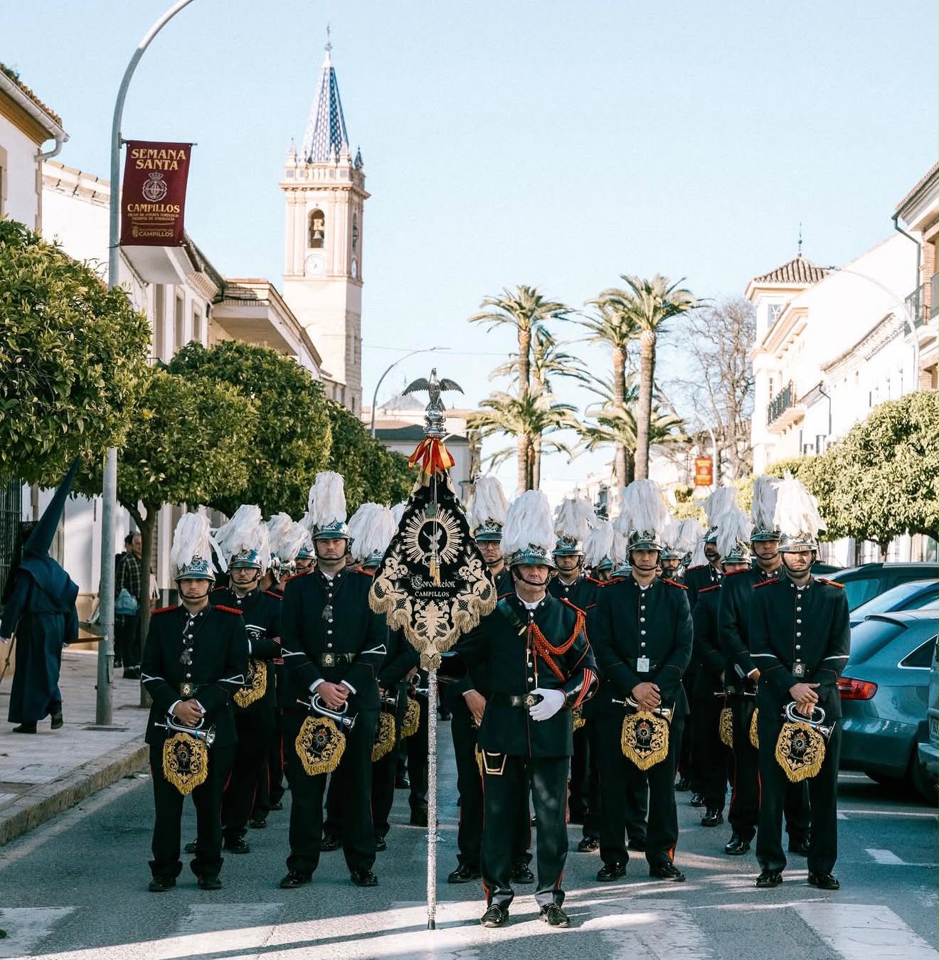 La Banda de Cornetas y Tambores Coronación formada en Campillos, con su banderín y la torre de la iglesia al fondo