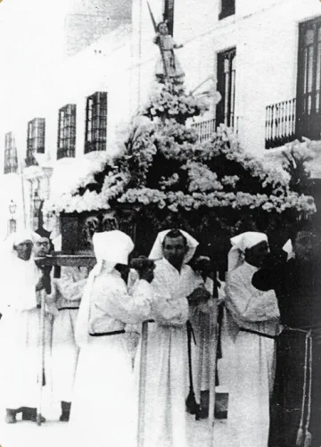 Bearers dressed in white carry a flower-covered Pollinica trono through the street, in a historic photograph.