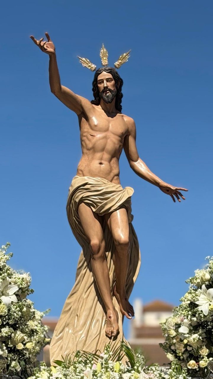 El Santísimo Cristo Resucitado con el brazo alzado, sobre el cielo azul y entre flores blancas