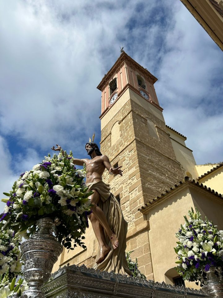 El Santísimo Cristo Resucitado en su trono, entre flores blancas y moradas, ante la torre de la Iglesia de Santa María del Reposo