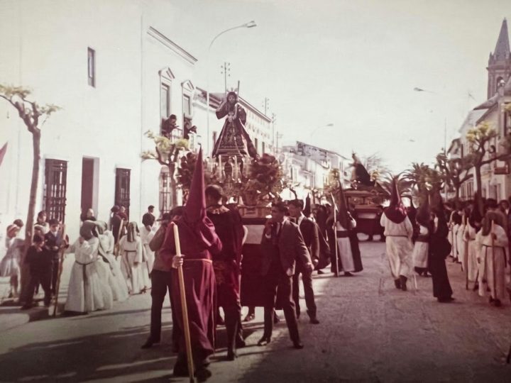 El trono del Dulce Nombre de Jesús por las calles de Campillos entre los nazarenos de túnica grana y blanca, en una fotografía antigua en color
