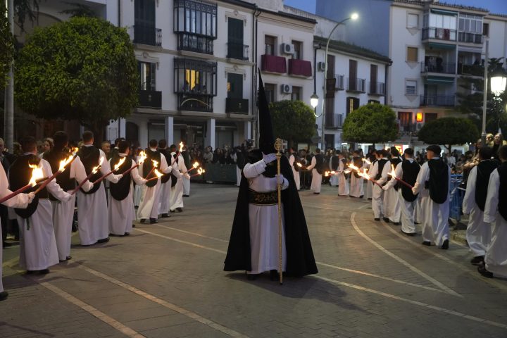 Un nazareno de capirote negro con vara dorada entre dos filas de hermanos con hachones encendidos