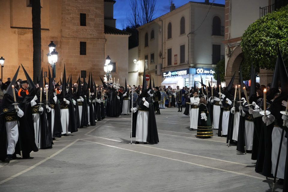Nazarenos de la Vera+Cruz con túnica blanca, capa y capirote negros y cirios encendidos, en dos filas al anochecer