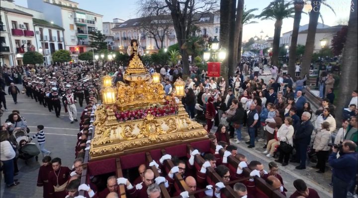 El trono del Dulce Nombre de Jesús a hombros de sus portadores de grana, al atardecer, seguido por la banda de música
